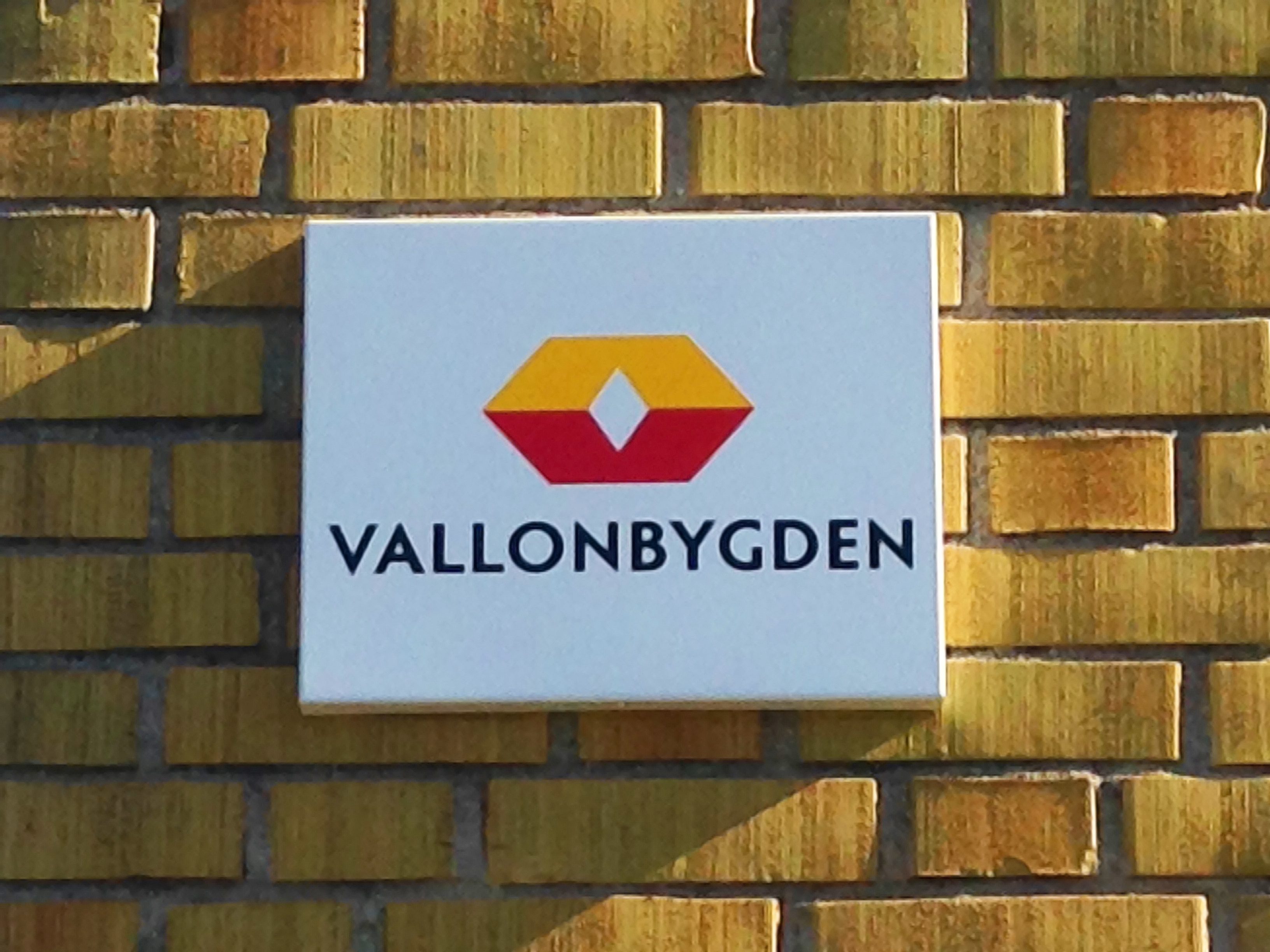 Bilden visar en gul tegelfasad med en skylt med Vallonbygden ABs logotyp på.